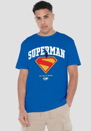 Young man wearing a blue Superman t-shirt with red and yellow logo and text "LAST SON OF KRYPTON," beige cargo shorts, gray background.