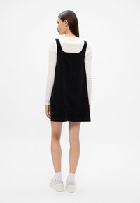 Μαύρο φόρεμα από κοτλέ με φαρδιές τιράντες και ίσια γραμμή, φορεμένο πάνω από λευκό μπλουζάκι με μακριά μανίκια, συνδυασμένο με λευκά sneakers.