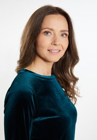 Zöld bársony felső magas nyakkivágással, testhez simuló fazonnal, hosszú ujjakkal és sima textúrával, fehér háttér előtt bemutatva.