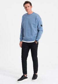 Ljusblå sweatshirt med rund hals och långa ärmar, i kombination med svarta tapered byxor och svarta sneakers. Enkelt design.