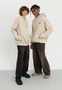Beige hoodies with drawstrings and front pockets, paired with dark wide-leg jeans. Black platform sneakers and light-colored clog-style shoes.