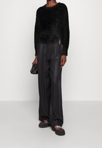 Person som bär en mjuk svart tröja, lösa svarta vida byxor, bruna vandringsskor och håller en liten svart handväska.