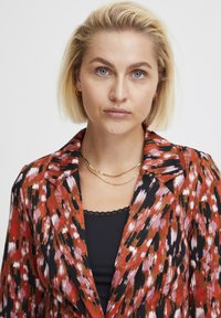 Színes mintás blazer piros, fekete és rózsaszín árnyalatokkal; sima anyagból készült, kihajtható gallérral és réteges dizájnnal.