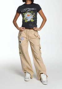 Mujer vestida con una camiseta gráfica negra con flores, pantalones cargo beiges con parches y zapatillas blancas, de pie con las manos sosteniendo el dobladillo de su camiseta.