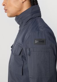 Prim-plan al unei persoane îmbrăcate cu o jachetă albastră închisă, cu guler înalt, având un buzunar cu fermoar și un patch cu logo-ul "Schott NYC" pe mânecă.