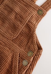 Salopette in velluto a coste marrone con scanalature verticali, caratterizzata da una tasca sul petto e tre bottoni in ottone etichettati "NEXT". Tessuto morbido con una tonalità calda.