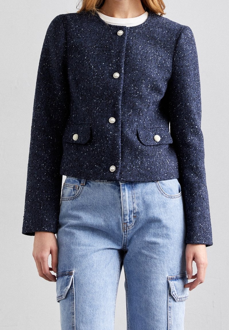 Femme portant une veste texturée bleu marine avec des boutons en perle et un jean cargo bleu clair sur un fond uni.