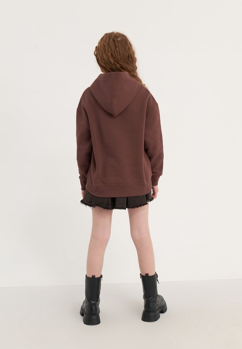 Jeune personne aux cheveux bouclés portant un sweat à capuche marron, une jupe foncée effilochée et des bottes noires montant jusqu’au milieu du mollet, debout devant un fond uni.