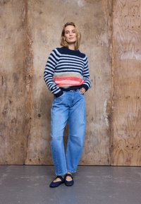 Pull en maille rayé avec nuances de bleu marine, gris et corail, associé à un jean taille haute bleu clair et des ballerines bleu foncé sur un fond uni.