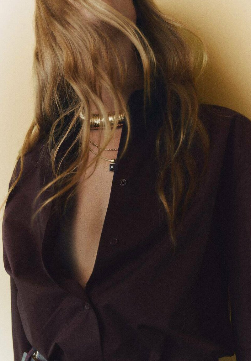 Camisa burdeos abotonada con un escote profundo, hecha de una tela suave. Collares de oro en capas añaden acentos metálicos.
