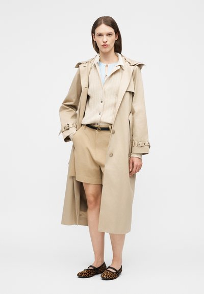 Femme portant un trench beige, un short beige, un pull léger et des ballerines imprimé léopard, debout devant un fond clair uni.