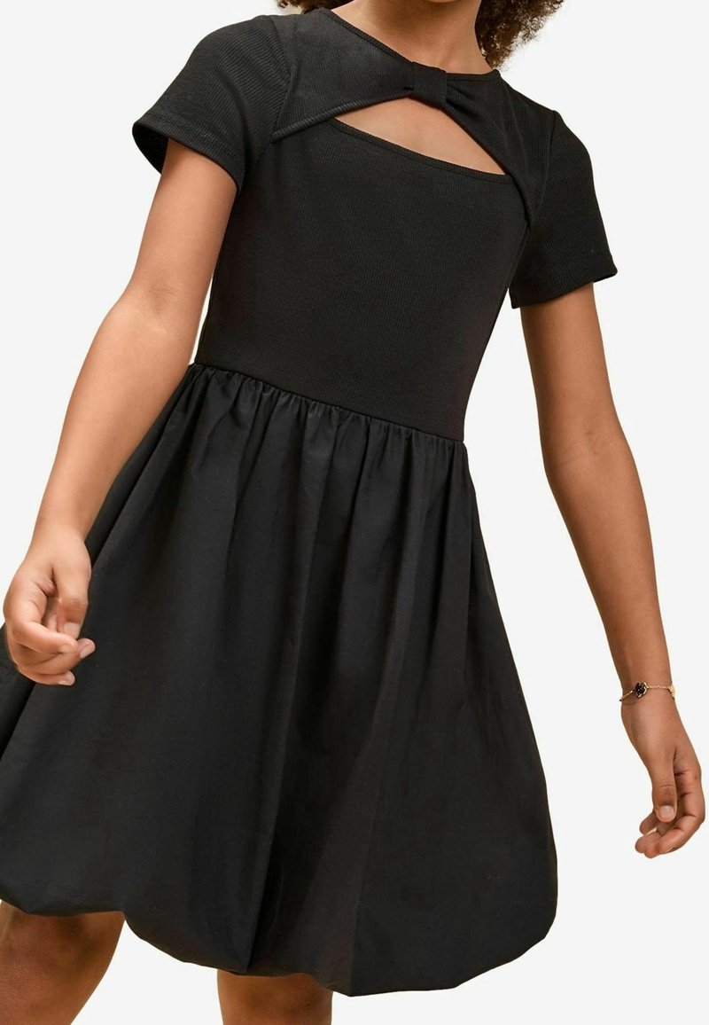 Fille portant une robe noire à manches courtes avec une jupe froncée et un décolleté carré avec un petit nœud.