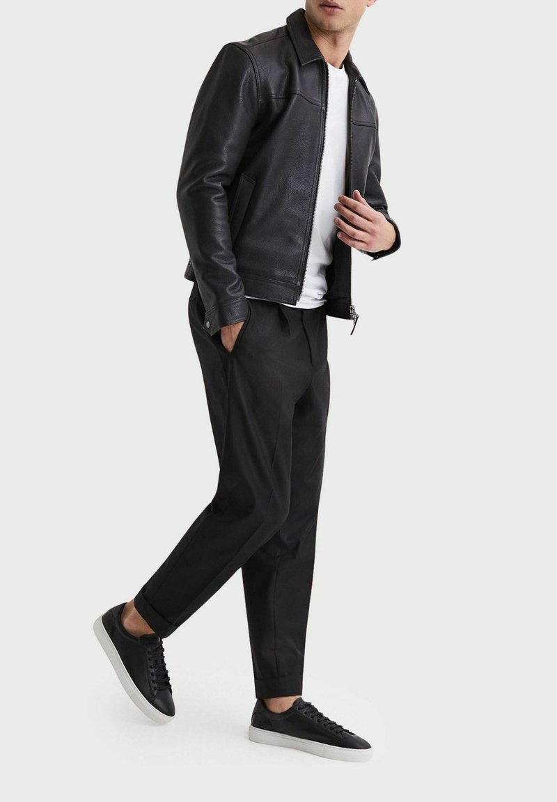 Schwarze Lederjacke mit glatter Textur, weißes T-Shirt, schwarze Hose mit lockerer Passform und schwarze Sneakers mit weißer Sohle.