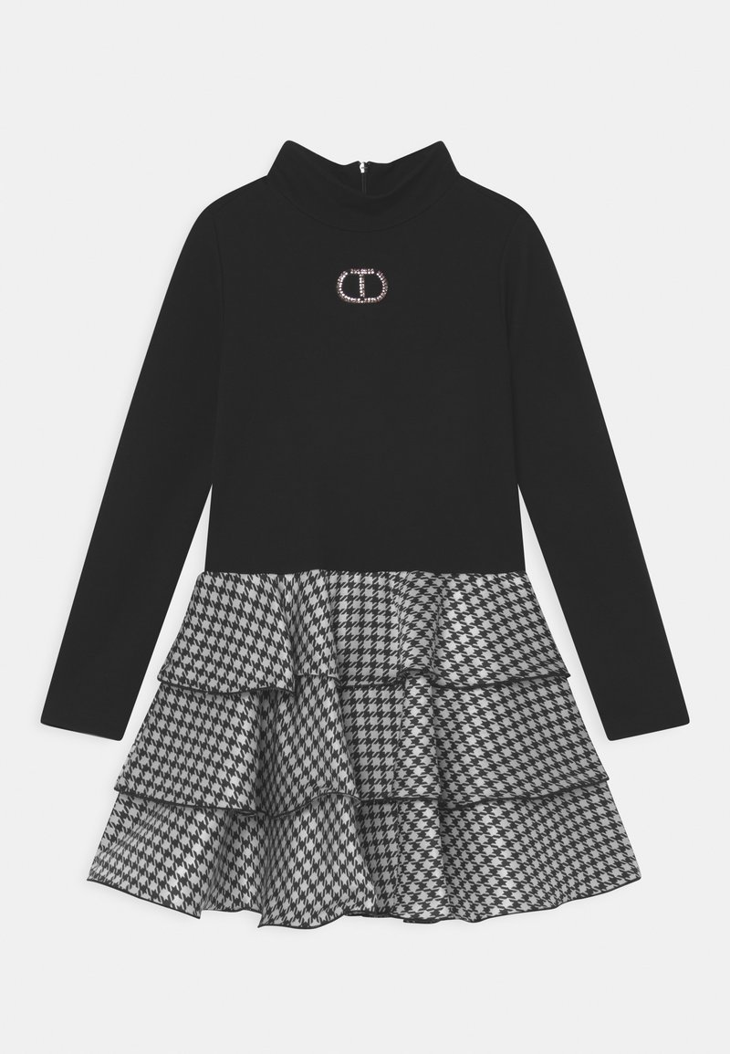 Robe noire à manches longues avec col cheminée, comprenant une jupe à volants à motif pied-de-poule gris et un logo décoratif sur la poitrine.