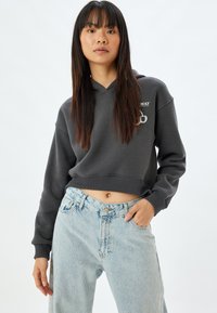 Sudadera gris oscuro ajustada con un bolsillo frontal y un logo, combinada con unos pantalones vaqueros de tiro alto en azul claro con patrones de diamantes y adornos.