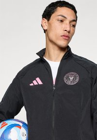 Μαύρο αθλητικό μπουφάν με ροζ ρίγες Adidas και λογότυπο Miami. Διαθέτει φερμουάρ και υφασμάτινη υφή. Κρατάει μια πολύχρωμη ποδοσφαιρική μπάλα.