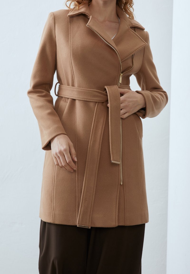 Manteau en mélange de laine couleur chameau avec une coupe ajustée, fermeture zippée, ceinture à nouer à la taille et manches longues. Texture lisse, sans motif.