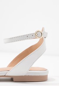 Białe skórzane sandały z niską brązową podeszwą, z cienkim paskiem na kostkę z złotą klamrą i przeszyciami wzdłuż krawędzi.
