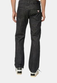 Jeans di denim scuro con una vestibilità dritta, caratterizzati da una toppa a forma triangolare e tasche posteriori. Il tessuto presenta una texture sottile e una leggera lucentezza.