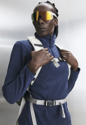 Άτομο που φοράει ναυτικό μπλε φλις με φερμουάρ, ανακλαστικά αθλητικά γυαλιά ηλίου και λευκό σακίδιο με ιμάντες στο στήθος και τη μέση, κοιτάζει ελαφρώς προς τα πάνω και στο πλάι.