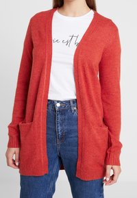 Röd lång kofta av mjukt stickat material med ribbade ärmslut och två framfickor, parat med en vit skjorta och högmidjade denim jeans.