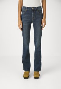 Jeans in denim blu scuro con design a gamba dritta, dotati di tasche e accessori metallici, abbinati a scarpe con piattaforma gialle.
