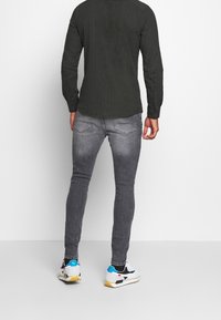 Donkergroen shirt met lange mouwen, grijze slim-fit jeans en witte sneakers met blauwe en gele accenten, gezien van achteren.