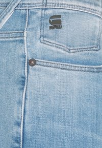 Jeans de mezclilla azul claro con un acabado texturizado. Presenta un parche de logo negro en el bolsillo trasero y un botón de metal con un ligero brillo.
