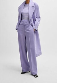 Lila trenchcoat med breda slag, över en ljusrandig topp och högmidjade, veckade byxor. Innehåller svarta skor.