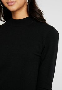 Pull en maille noir avec un col montant et des détails côtelés. Texture lisse et coupe ajustée, mettant en valeur l'épaule et le col.