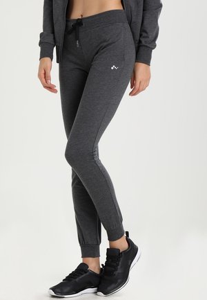 Nainen, jolla on päällään tummanharmaat vetoketjulliset hupulliset housut, joissa on nyörivyötärö ja resorit nilkoissa, mustat lenkkarit sekä vastaava vetoketjullinen takki.