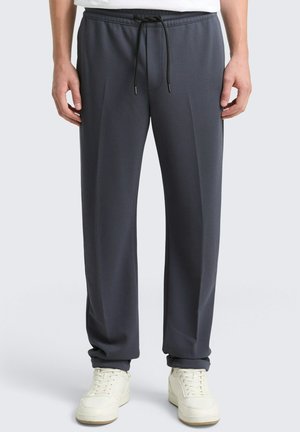 Mann trägt dunkelgraue Jogginghose mit Kordelzug und weiße Sneakers, steht vor einem einfarbigen hellen Hintergrund.