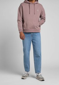 Pink hoodie with a front pocket, drawstring hood, and small logo patch, paired with light blue straight-leg jeans and gray sneakers.