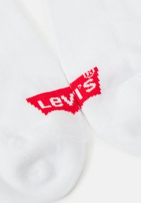 Fehér zoknik, amelyek domború piros "LEVI'S" logóval rendelkeznek, egy stilizált háromszög mintában a szárakon. Simább textúra, kötött anyag.