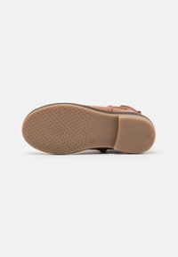 Sandal de color marrón con suela de goma texturizada, correa ajustable y diseño minimalista. El material suave y el color claro mejoran la comodidad.