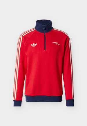 Czerwona długa bluza sportowa z długim rękawem, granatowym kołnierzem i mankietami, białe paski na rękawach, logo Adidas na lewej piersi oraz herb działu na prawej piersi.