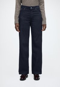 Temno modre široke kavbojke z visokim pasom, iz jeansa, s sprednjimi žepi in nežnimi šivi. Kombinirane s črnimi čevlji.