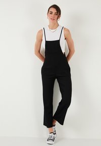 Salopette noire avec un corsage ajusté et des jambes larges, portée par-dessus un débardeur blanc. La tenue présente des bretelles réglables et des poches latérales.