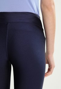 Marina träningsleggings med hög, slät midjeband och en figurnära design, tillverkade av ett glänsande, stretchigt material. Närbild av bakifrån.