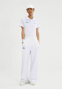 La camisa polo blanca y los pantalones de pierna ancha presentan detalles y marcas en azul. El atuendo se completa con una gorra blanca a juego y muñequeras.