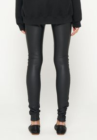 Leggings in ecopelle nera con un design aderente, texture liscia e taglio alla caviglia, abbinati a scarpe basse nere e una maglia scura.