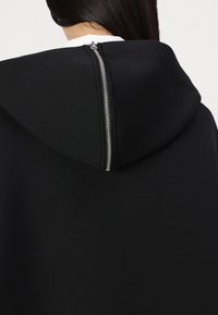 Sweat à capuche noir avec un zip distinct au dos, fabriqué dans un tissu doux et texturé avec un col structuré.