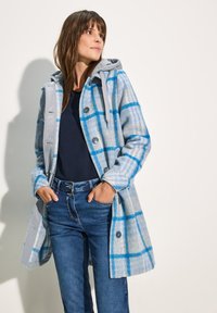 Cappotto a quadri grigio e blu con cappuccio, dotato di grandi bottoni e una texture morbida. Indossato sopra un top navy e jeans in denim.
