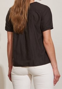 Camisetta nera a maniche corte con tessuto testurizzato, dettaglio arricciato sul retro e polsini elasticizzati, abbinata a jeans bianchi aderenti.
