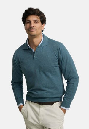 Hombre con pelo oscuro que lleva una camisa polo azul de manga larga sobre una camisa de cuello a rayas y pantalones beige claros, con las manos en los bolsillos.