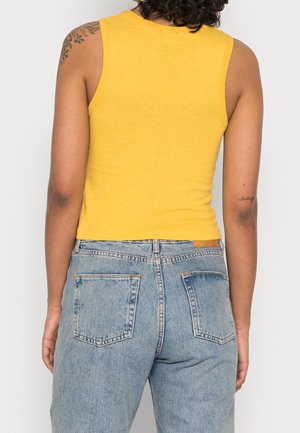 Persona con camiseta sin mangas amarilla y vaqueros de tiro alto y lavado claro, vista desde atrás con los brazos relajados a los lados.