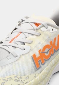 Chaussure de course gris clair avec tige en mesh, marquage orange, semelle texturée et lacets. Présente un motif de rayures subtil et des accents en caoutchouc durable.