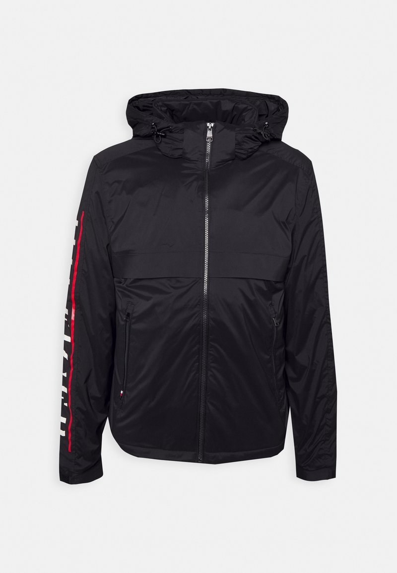 Veste noire à capuche avec fermeture éclair à l'avant, deux poches latérales zippées, et un design rayé rouge et blanc le long de la manche gauche.