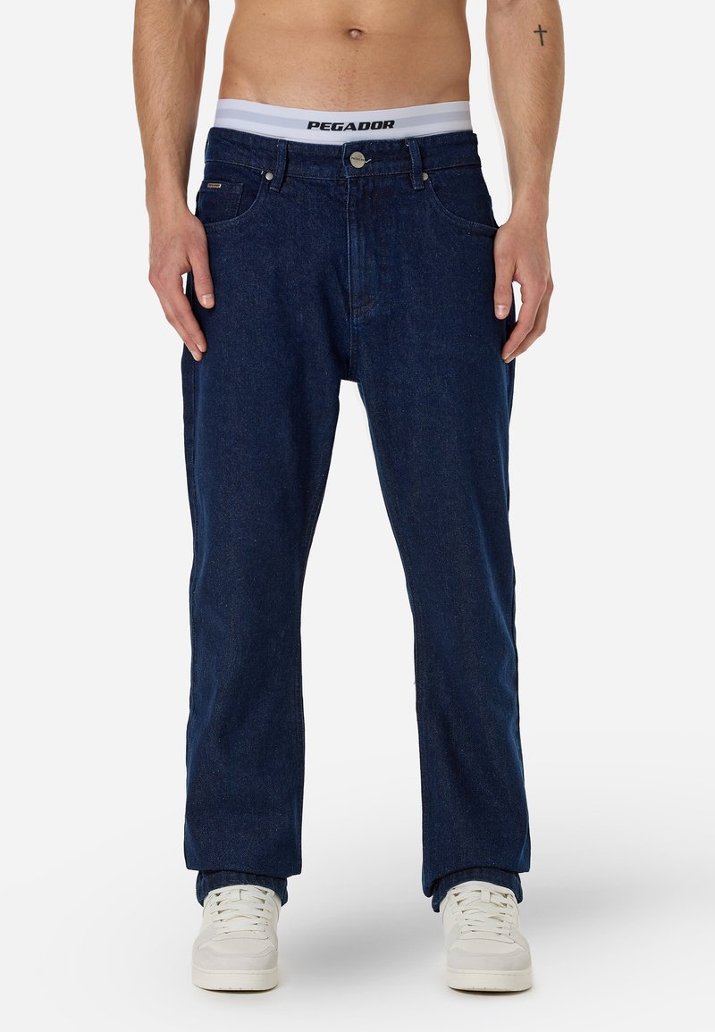 Σκούρο μπλε τζιν παντελόνι με ίσια γραμμή, που διαθέτει μπροστινές τσέπες, κλείσιμο με κουμπί και ορατή ζώνη με την ετικέτα "PEGADOR."