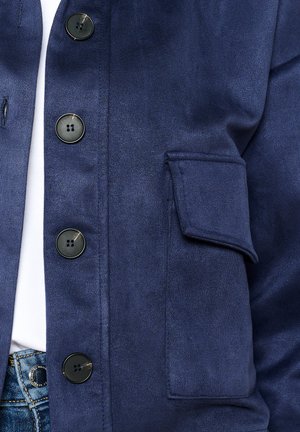 Tummansininen takki, joka on valmistettu sileästä kankaasta. Takissa on mustat napit, läppätasku ja hillitty kiilto, sekä nappikiinnitys edessä.